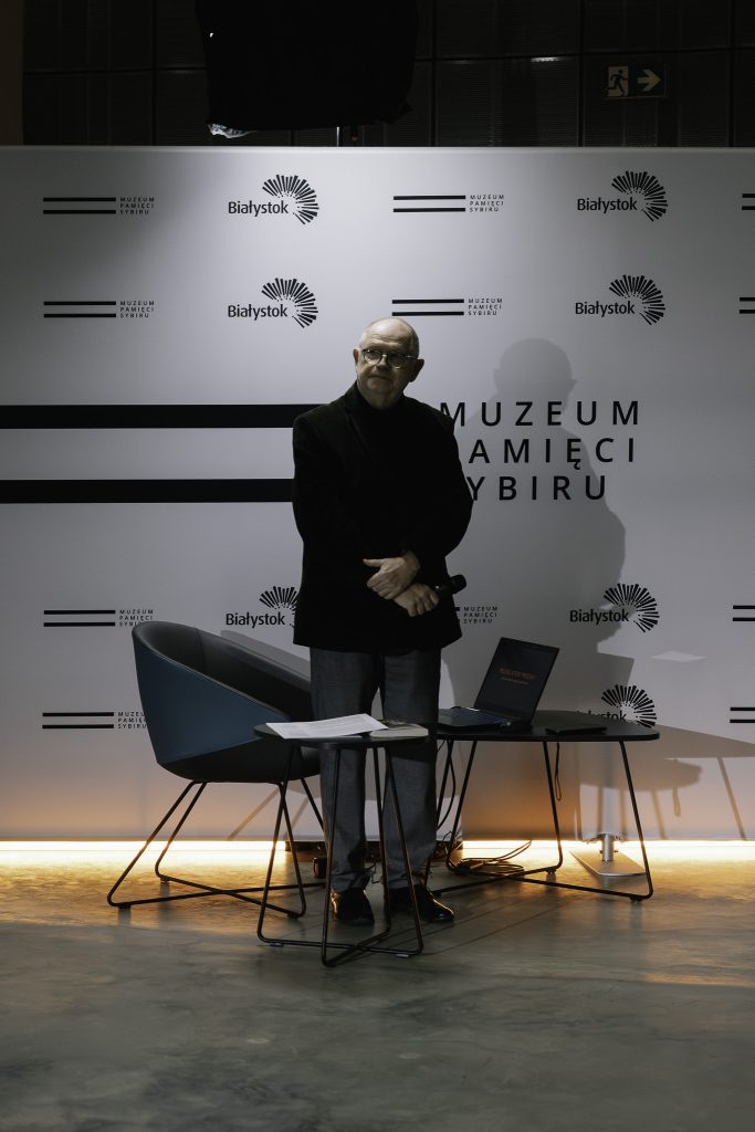 Krzysztof Renik stoi na tle logo Muzeum Pamięci Sybiru oraz logo Miasta Białystok. Ręce ma opuszczone, w jednej z rąk trzyma mikrofon. Spogląda w stronę ekranu z rzutnika. Obok niego stoi fotel oraz stolik na którym znajduje się laptop.