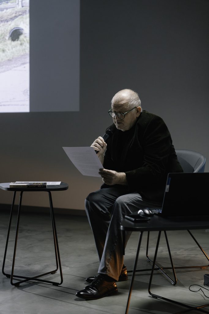 Krzysztof Renik siedzi na fotelu. W prawej ręce trzyma mikrofon. W lewej ręce trzyma kartkę z której właśnie czyta. Jest to starszy mężczyzna w okularach. Obok niego stoją dwa niewielkie stoliki. Na jednym z nich jest książka, na drugim jest laptop. W tle na ścianie widoczny jest fragment obrazu z rzutnika.