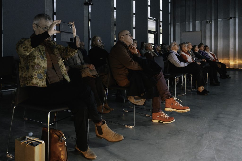 Grupa kilkunastu osób siedzi na krzesłach. W dużym skupieniu słuchają opowieści oraz oglądają wyświetlane zdjęcia.