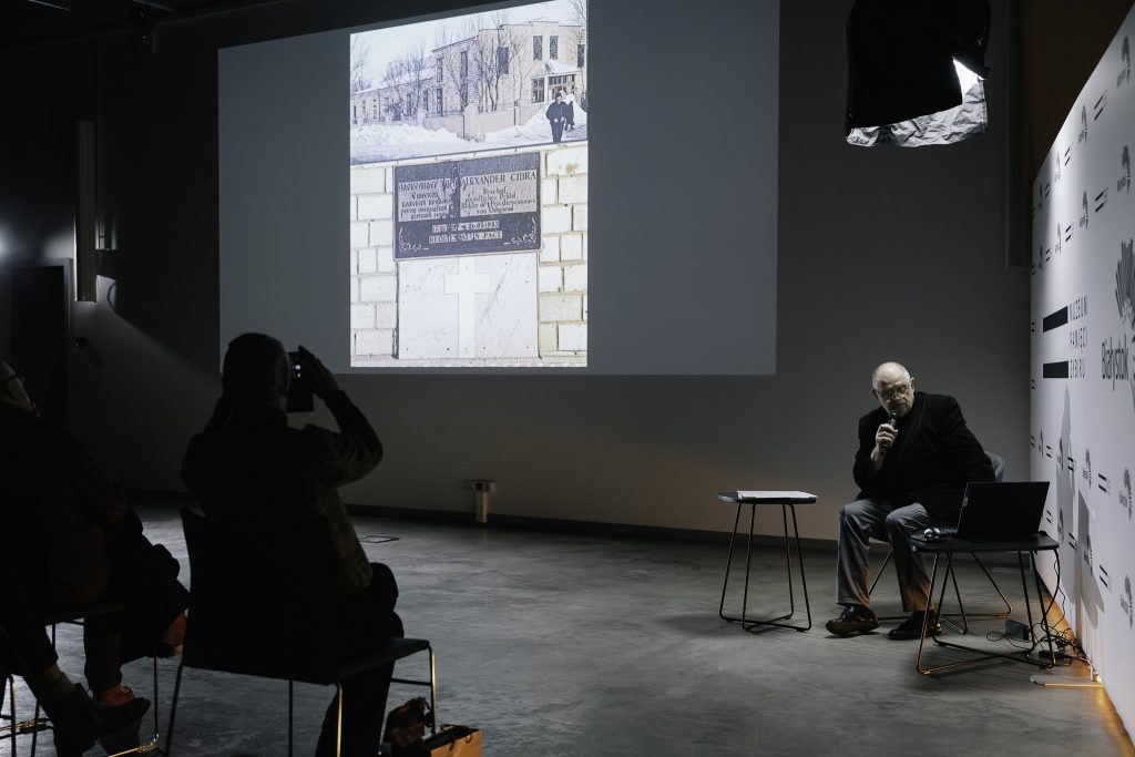 Krzysztof Renik opowiada o książce. Jest to starszy mężczyzna. Siedzi na fotelu. W ręku trzyma mikrofon. Spogląda na ekran laptopa. Na dużym ekranie z rzutnika jest wyświetlane jedno z zdjęć z podróży po terytorium byłego Związku Radzieckiego.