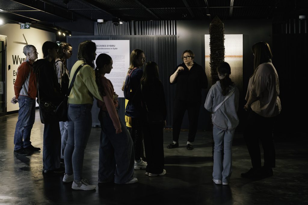 Grupa uczestników warsztatów jest na wystawie czasowej. Słucha wykładu Katarzyny Zabłockiej. Stoją w słabo oświetlonej sali, w tle widoczne eksponaty.