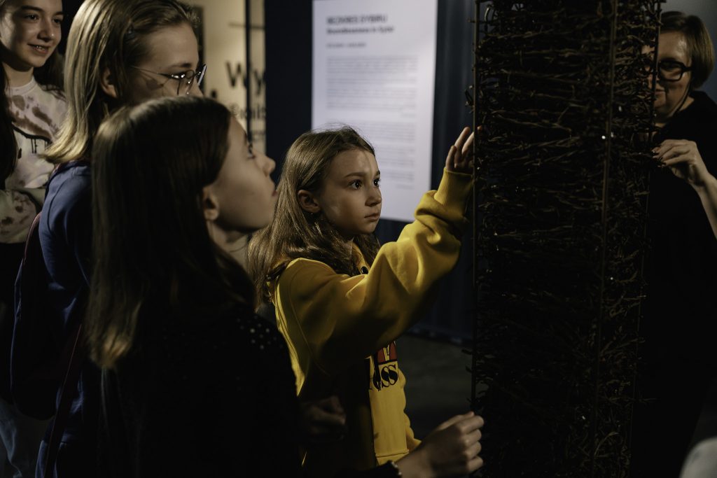 Grupa dzieci stoi przy jednym z eksponatów. Jest to słup z drutu kolczastego. Jedna z dziewczynek ostrożnie dotyka eksponatu.