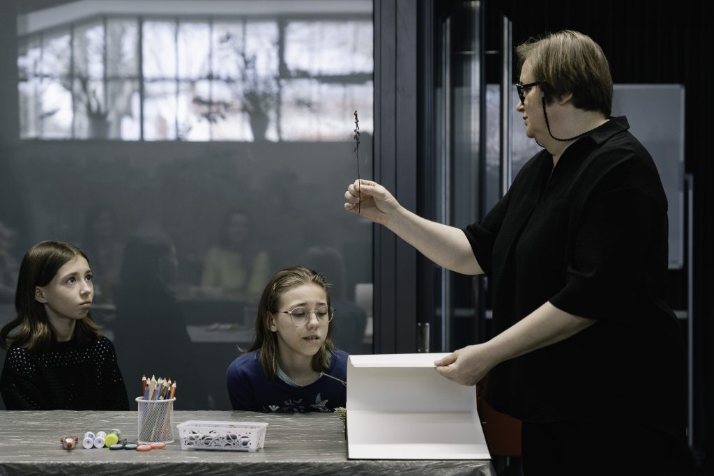 Katarzyna Zabłocka prowadzi warsztaty. Dwie dziewczynki siedzą przy stole i uważnie słuchają instrukcji.
