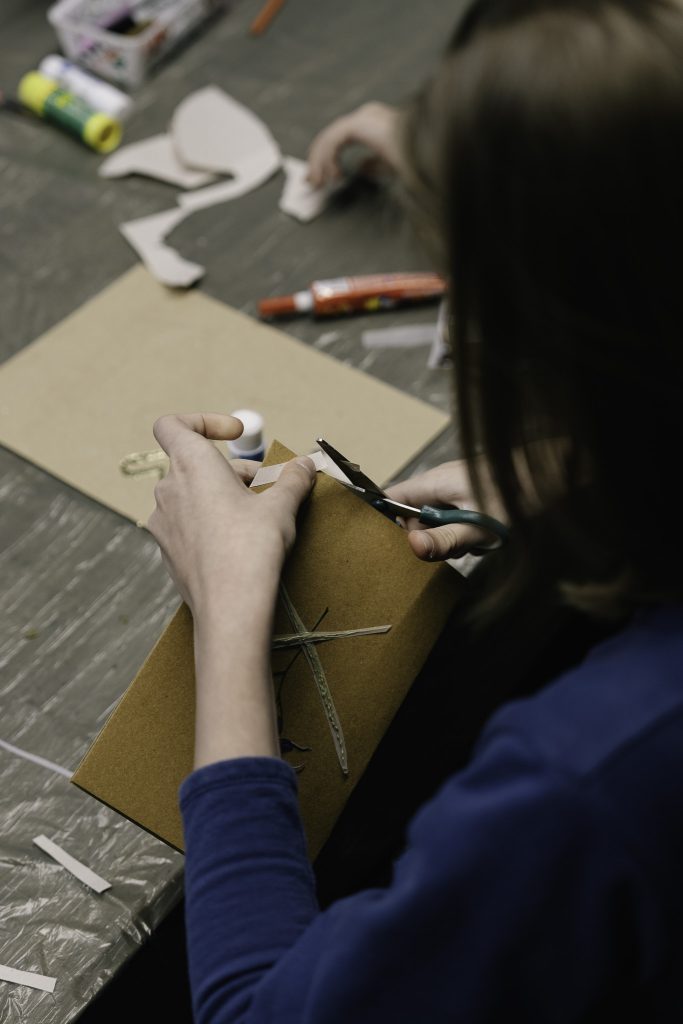 Młoda dziewczyna pilnie pracuje nad pocztówką dla Sybiraka. Trzyma w ręku małe nożyczki. Powstająca pocztówka ma brązową, skromną okładkę z naklejoną suszoną rośliną.