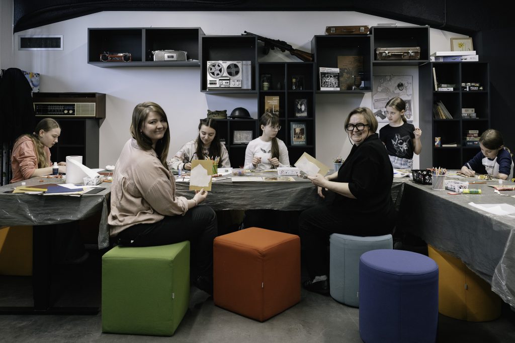 Młodzież pracuje na warsztatach. Na stołach leżą różne przybory biurowe, kleje, kredki, kartki papieru, nożyczki. Ich pracę nadzorują Ewelina Kamieńska oraz Katarzyna Zabłocka.