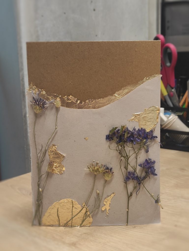 Na stole stoi jedna z gotowych pocztówki dla Sybiraka. Na pocztówce są naklejone suszone drobne roślinki przetykane złotymi wstawkami. Tło pocztówki to szary papier tekturowy.