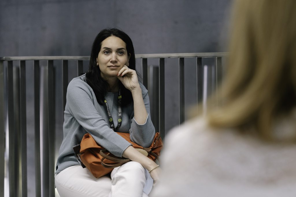 Kobieta w brązowych włosach, niebieskiej bluzce z pomarańczową torbą na kolanach słucha podczas oprowadzania po wystawie muzeum.